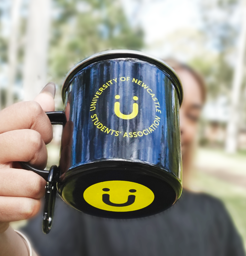 A close-up shot of a hand holding a black enamel mug with a yellow smiley face logo and the text 'UNIVERSITY OF NEWCASTLE STUDENTS ASSOCIATION' printed on it. The background shows a blurred outdoor scene with trees and a person.